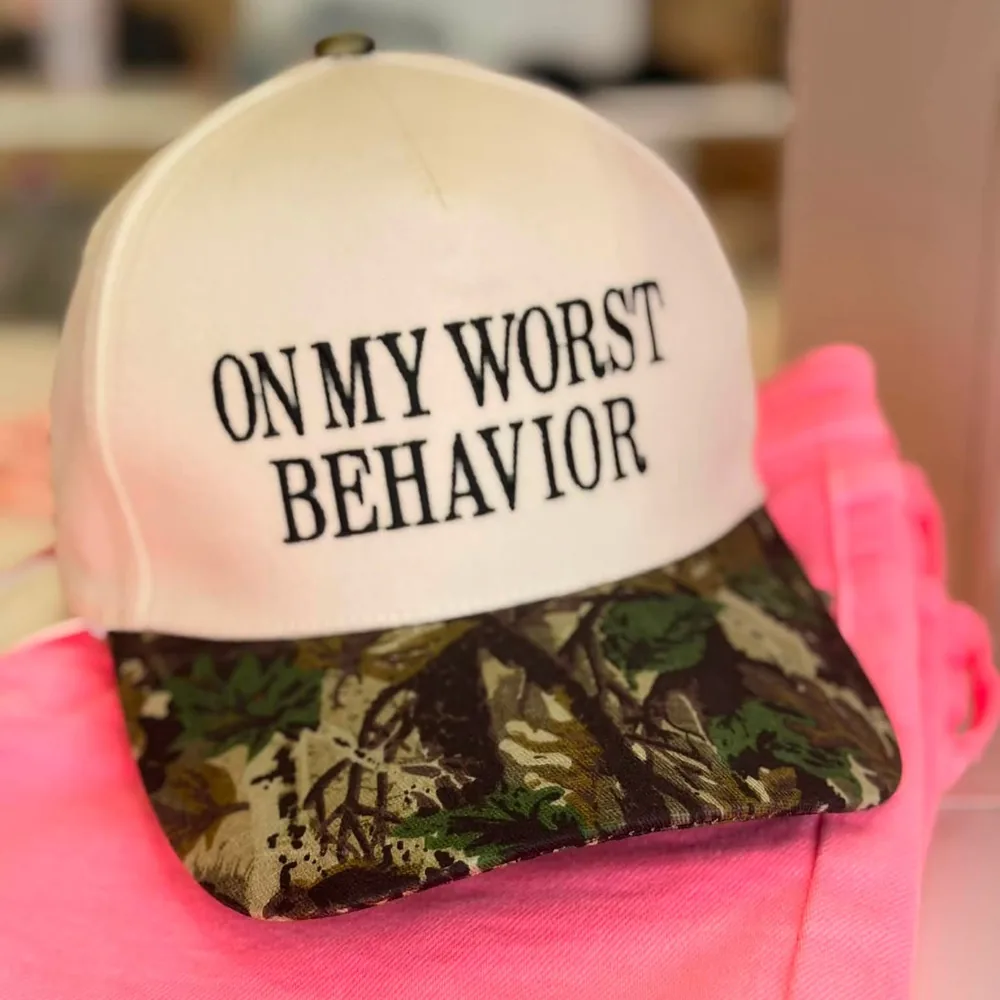 Cream trucker cap with a Realtree camouflage brim, embroidered in black block letters: On My Worst Behavior, resting on a hot pink garment.