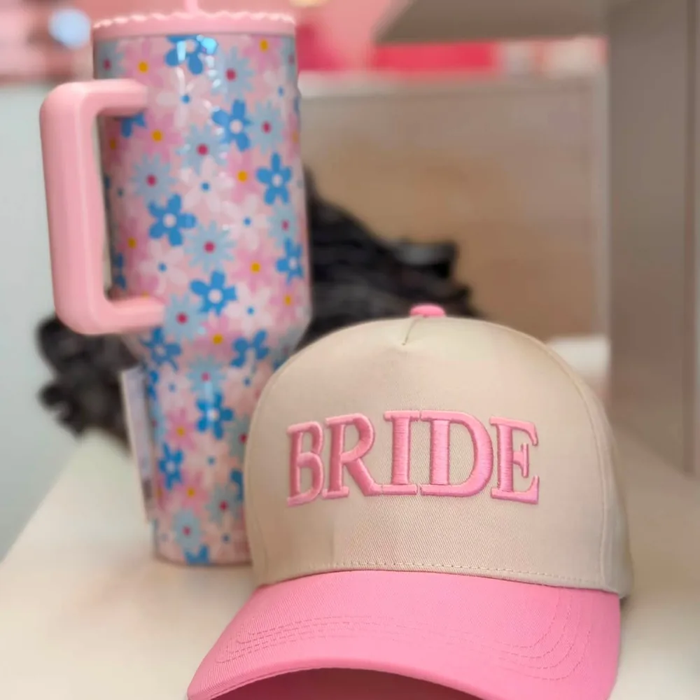 Cream trucker cap with a pink brim, embroidered BRIDE in raised pink block letters, next to a pink and blue daisy print tumbler.