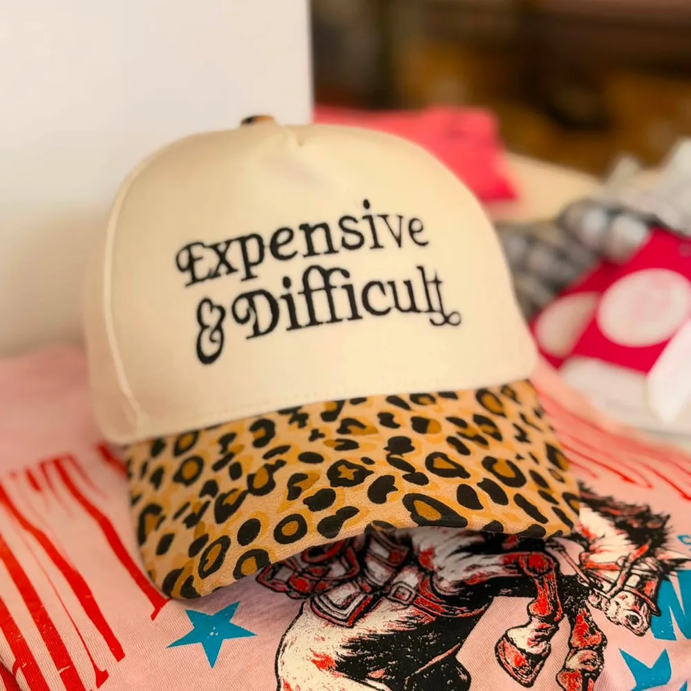 Cream trucker cap with a leopard print brim, embroidered in ornate black Victorian type: Expensive and Difficult. It rests on a pink rodeo tee.