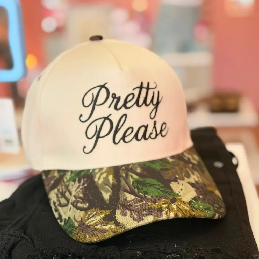 Blush trucker cap with a camouflage brim, embroidered in black script: Pretty Please, sitting on folded black distressed denim.